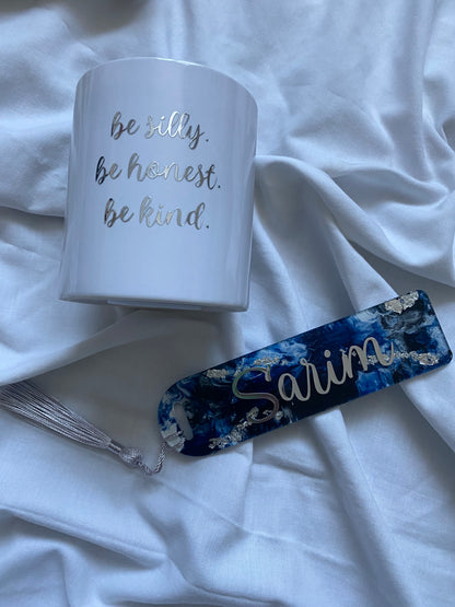 A white ceramic bookmark with gold text, alongside a blue and gold floral patterned bookmark, both with silver tassels.
